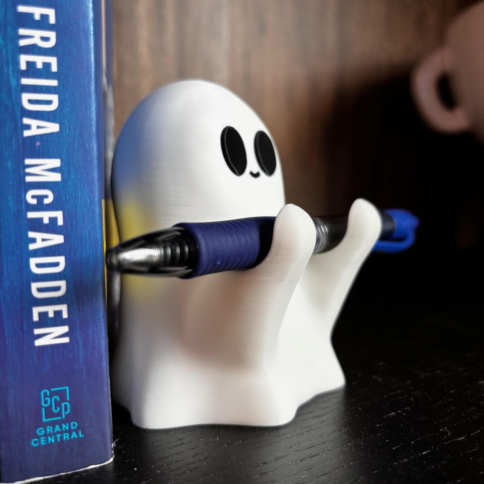 A cute white ghost-shaped pen holder grips a blue and black pen. Next to it, a blue book spine reads Freida McFadden in white letters. Both items rest on a dark wooden surface.