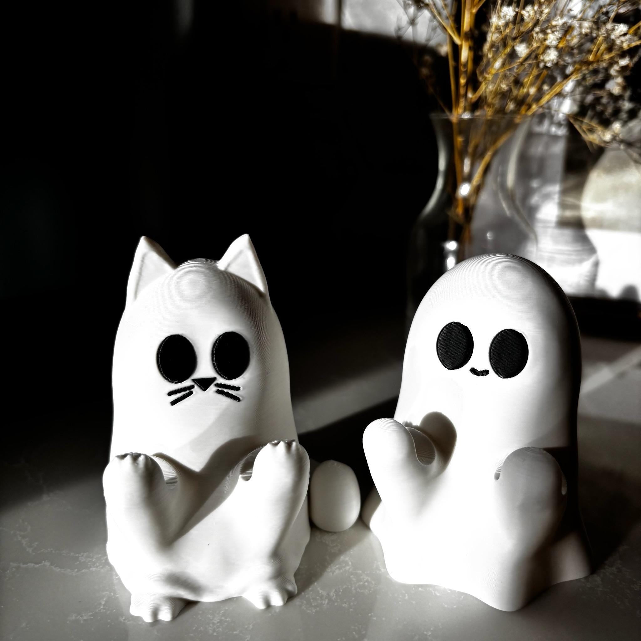 Two small white figurines sit on a surface: one shaped like a cat with round eyes and whiskers, and one shaped like a ghost with round eyes and a small smile. A vase with dried flowers is blurred in the background.
