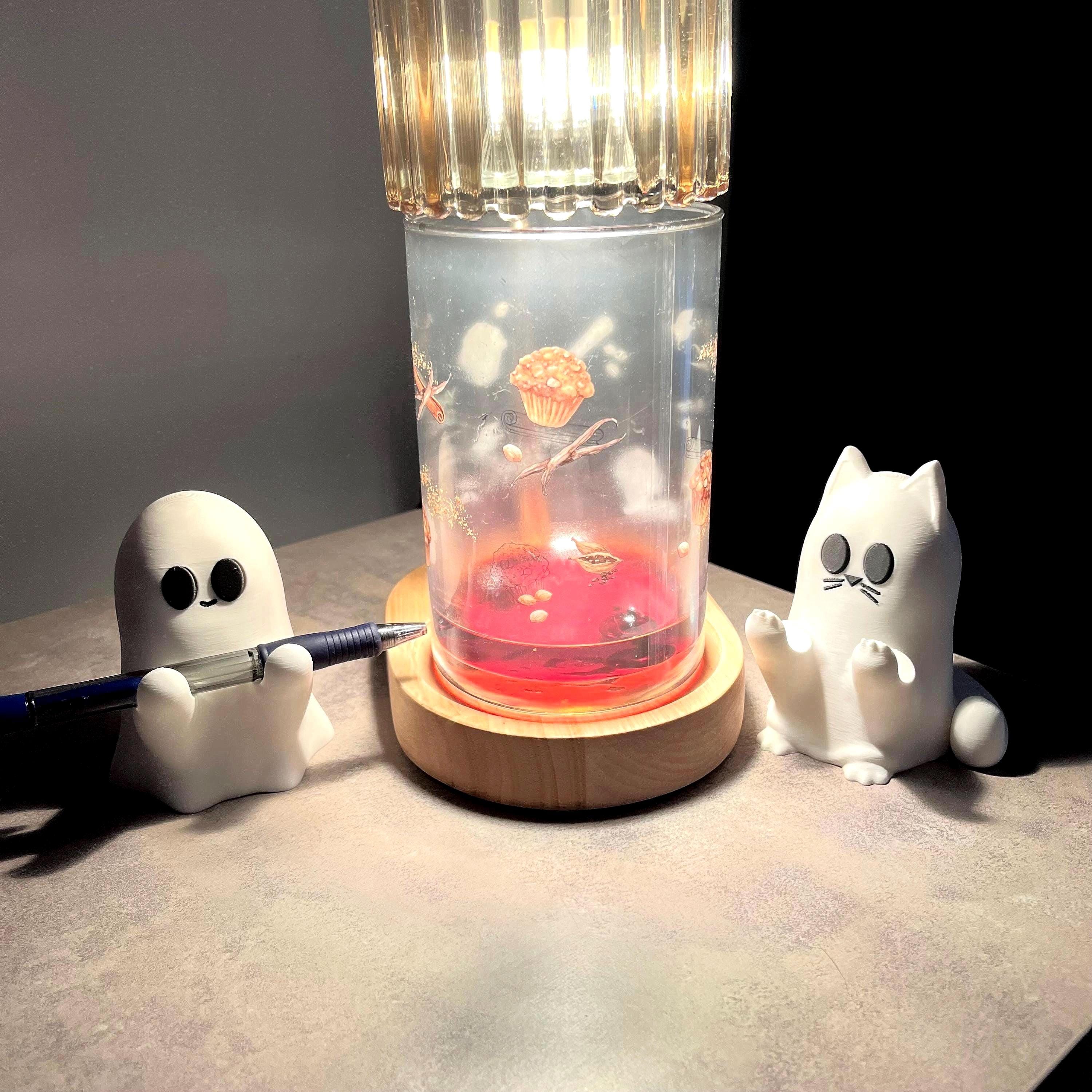Two small white figurines, one a ghost holding a pen and the other a cat, sit beside a lamp with a red liquid base, casting warm light on a flat surface in an otherwise dimly lit room.