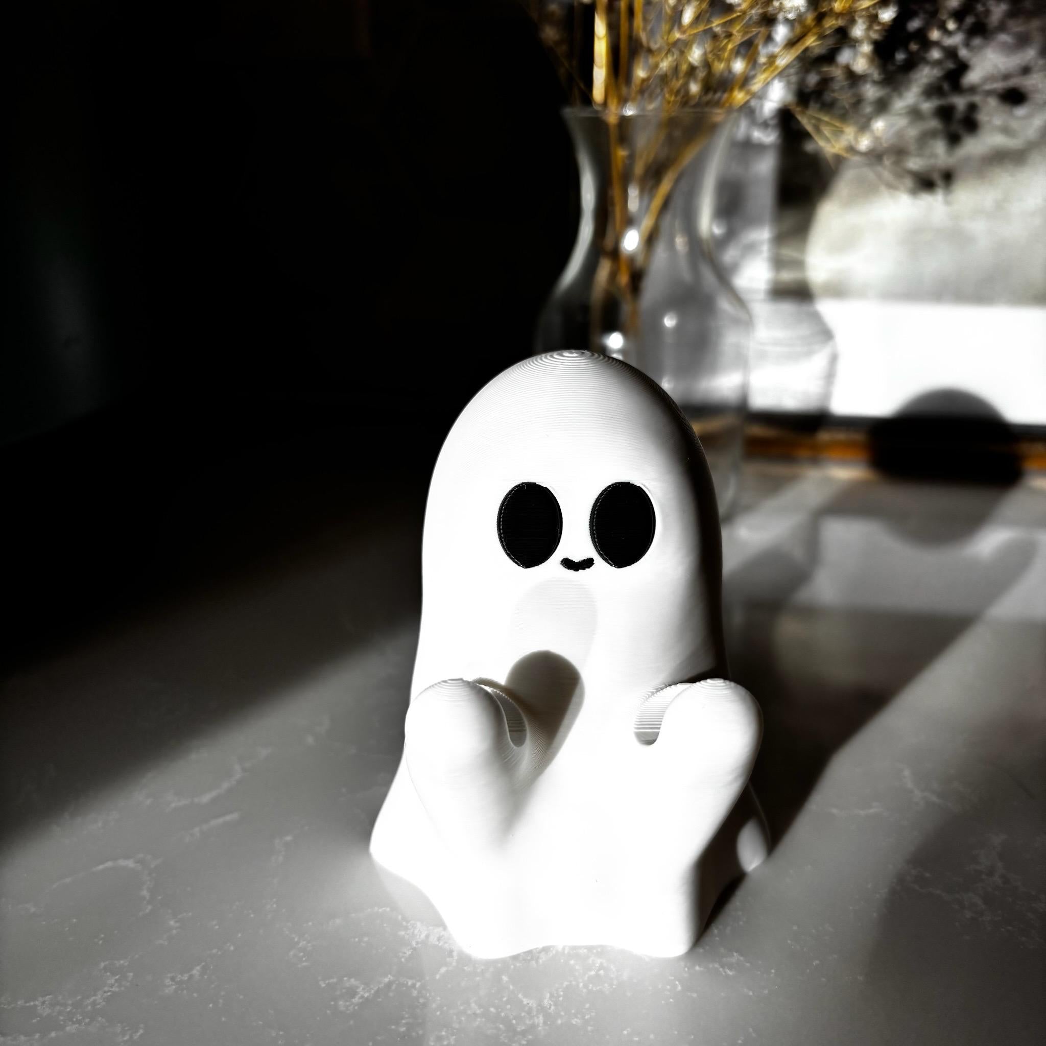 A small white ghost figurine with black eyes and a smile sits on a white surface, with sunlight casting shadows. Blurred glass vases and dried plants are visible in the background.