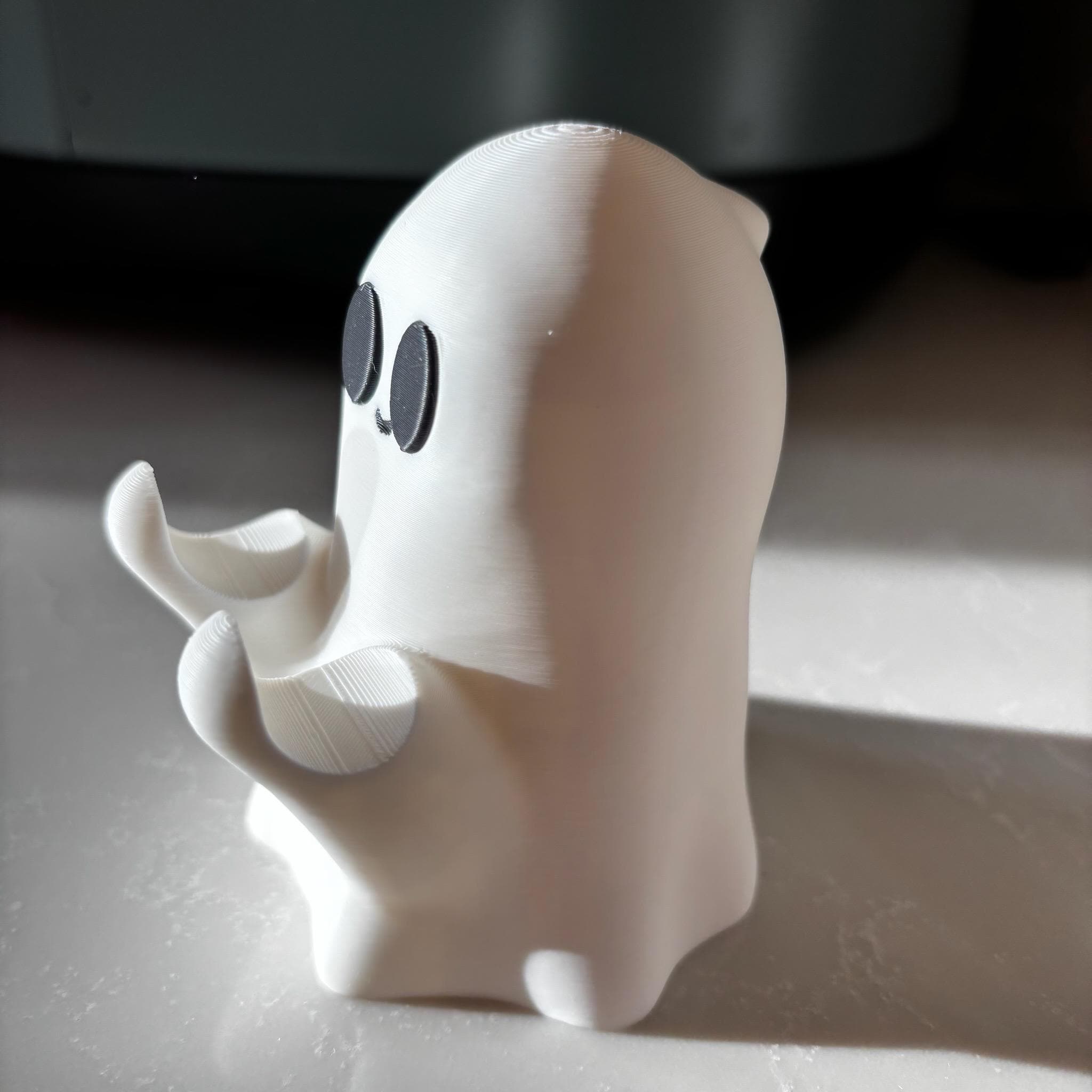 A small, white, 3D-printed ghost figurine with large black eyes and outstretched arms is standing on a sunlit surface.