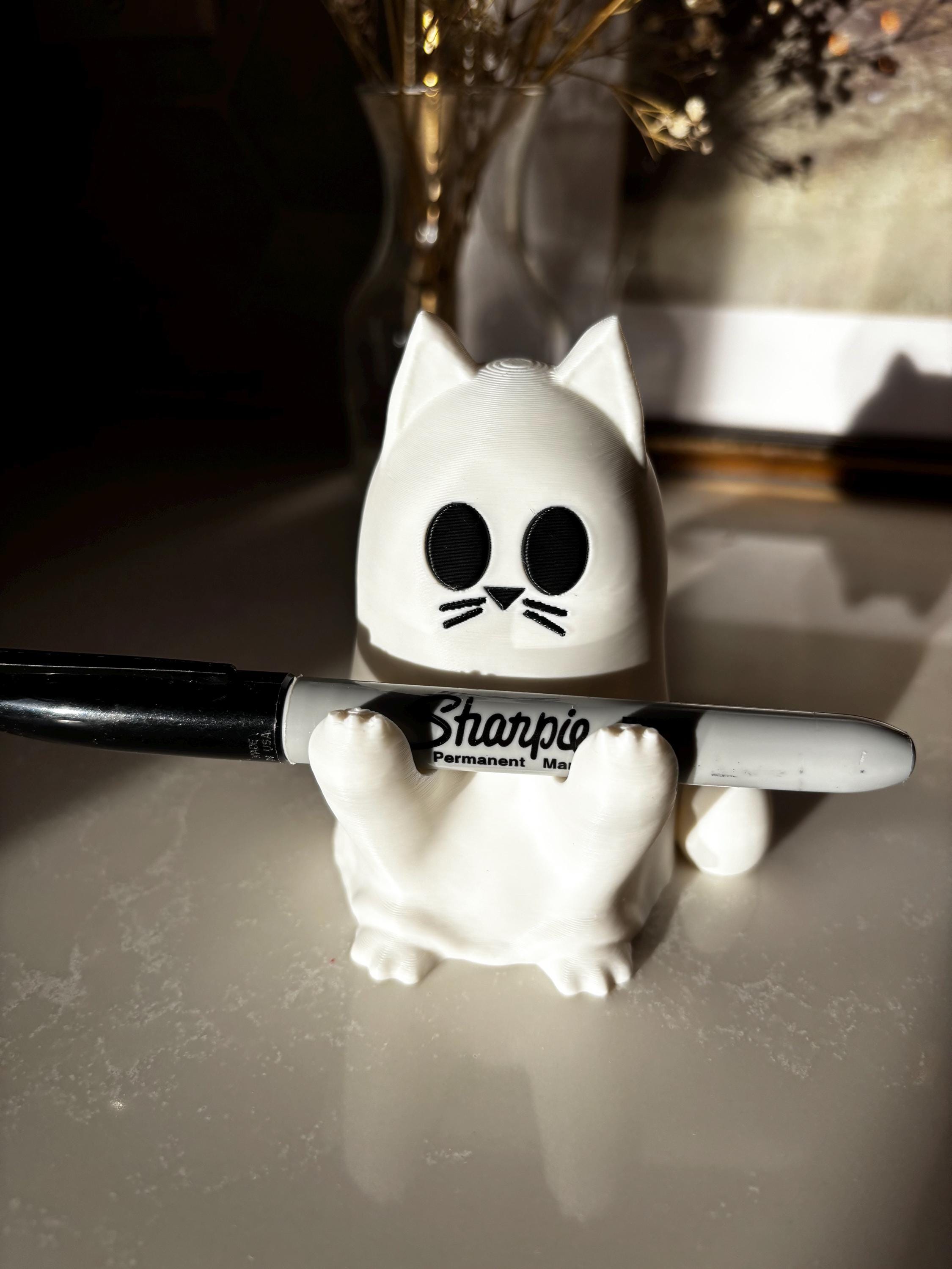 A white cat-shaped ghost with black eyes and whiskers is holding a black Sharpie permanent marker on a glossy surface, with a blurred background.