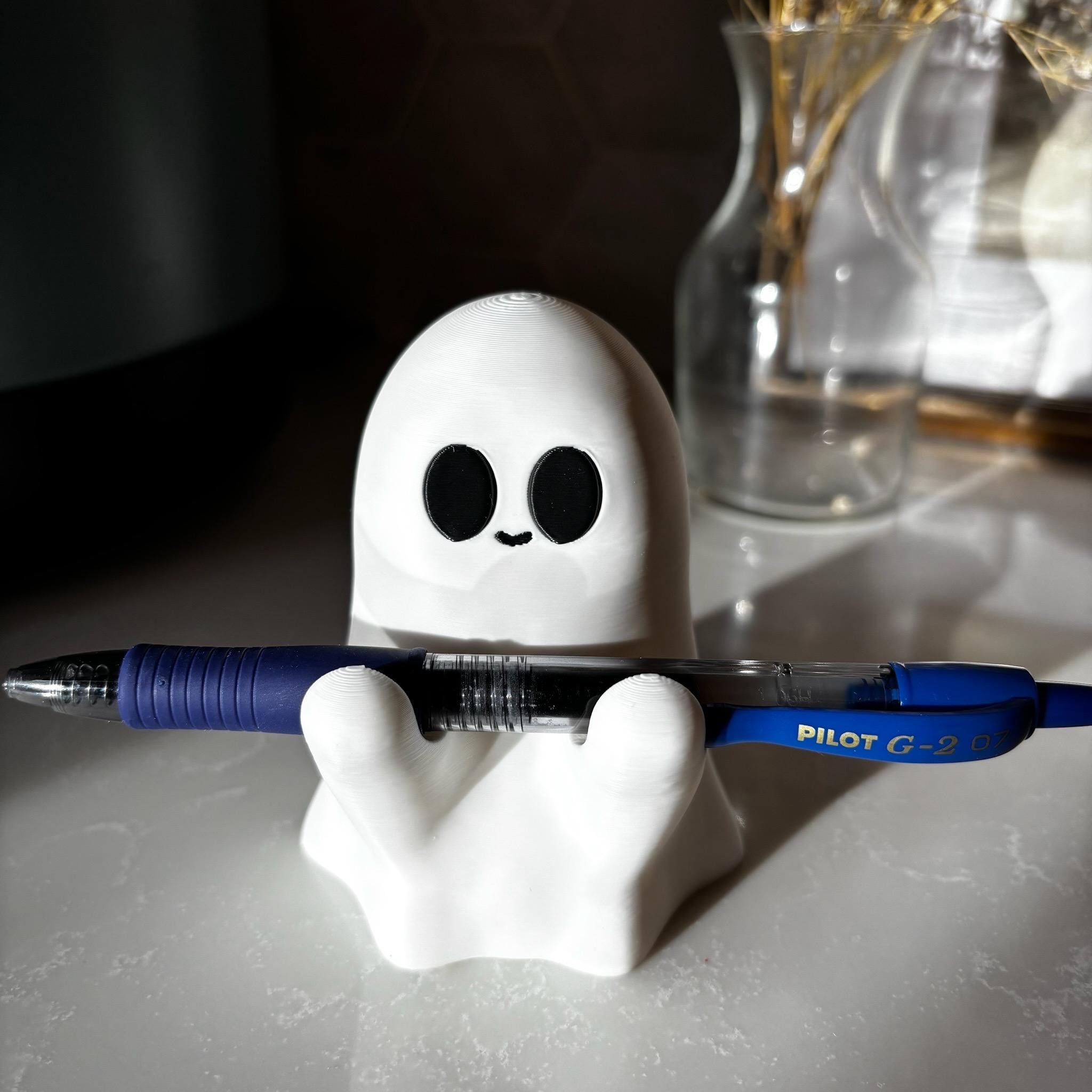 A small white ghost-shaped holder with black eyes is holding a blue Pilot G-2 pen on a countertop. In the background, there are glass vases with dried plants.
