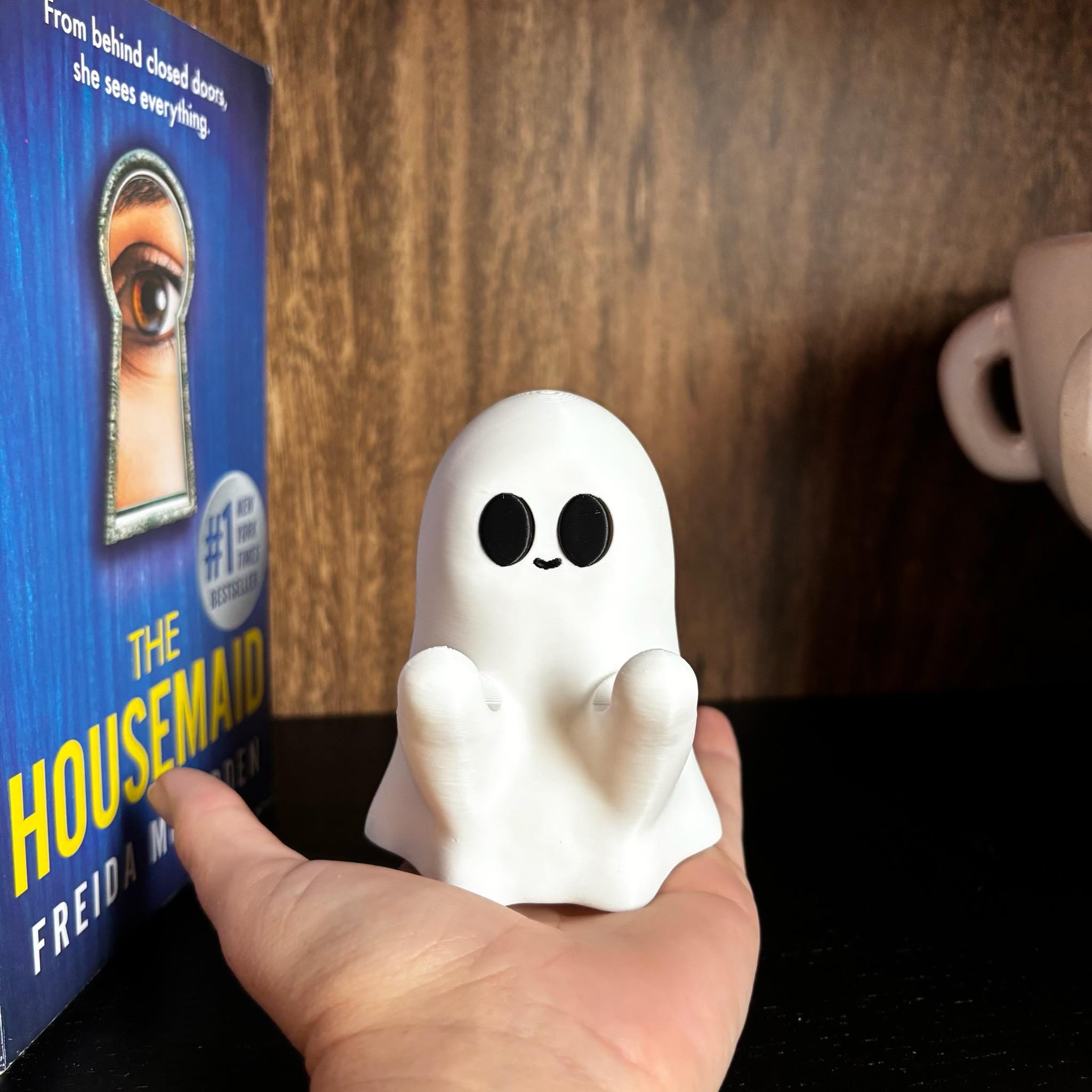 A hand holds a small, cute ghost figurine with big black eyes and a smile. To the left is a book titled The Housemaid by Freida McFadden, and to the right is part of a light-colored mug.