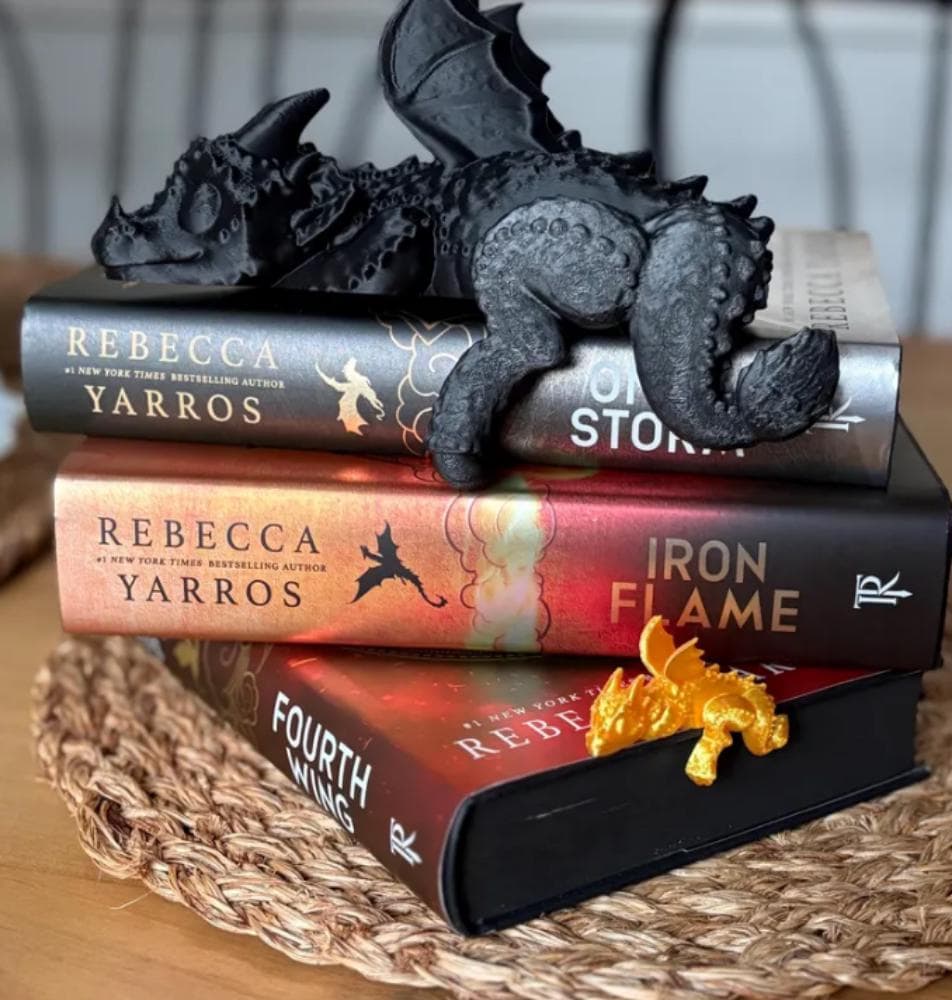Sleeping black 3D printed dragon figurine rests across stacked Fourth Wing books, with small golden dragon accent.