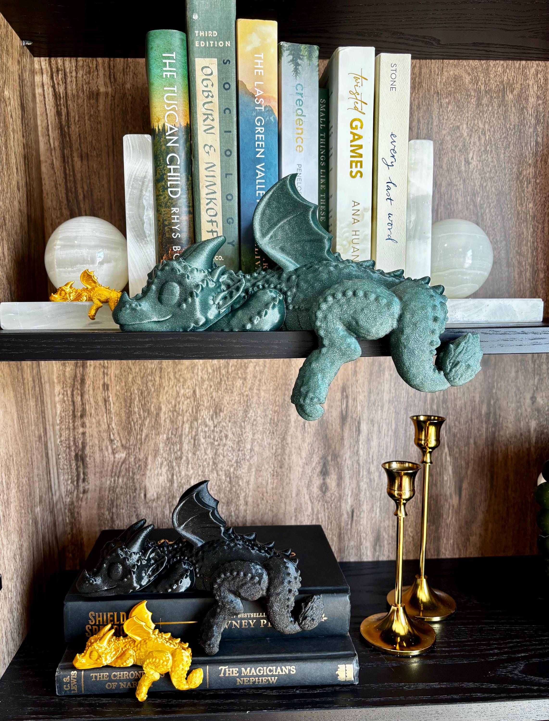Side view of family of fourth wing dragons, black sleeping dragon 3D print draped over book spines, wing tucked under chin – compact bookshelf ornament highlighting texture and silhouette for Google search. Gold silk dragon, glitter green dragon