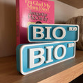 Two teal and white BIO BLVD bookshelf street signs, small stacked on large, in front of a book