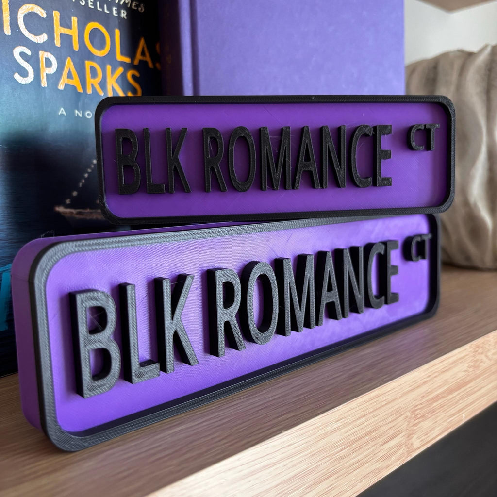 Purple and black BLK ROMANCE CT street signs, small stacked on large, on a bookshelf