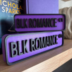 Purple and black BLK ROMANCE CT street signs, small stacked on large, on a bookshelf