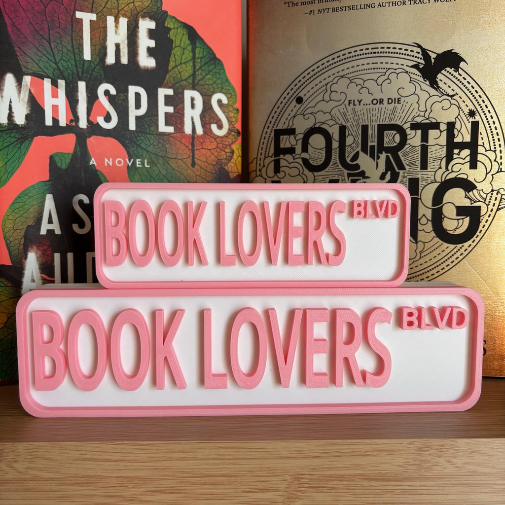 Pink and white BOOK LOVERS BLVD street signs, small stacked on large, in front of books