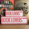 Pink and white BOOK LOVERS BLVD street signs, small stacked on large, in front of books