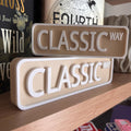 Beige and white CLASSIC WAY street signs, small stacked on large, on a bookshelf