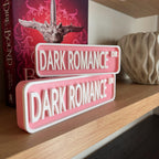 Pink and white DARK ROMANCE DR street signs, small stacked on large, in front of a book