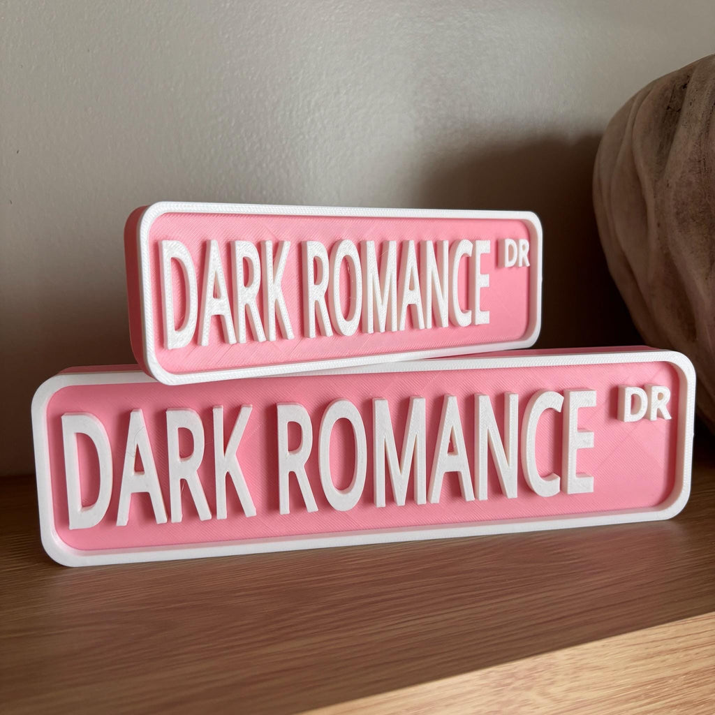 Small and large pink and white DARK ROMANCE DR street signs on a wooden shelf