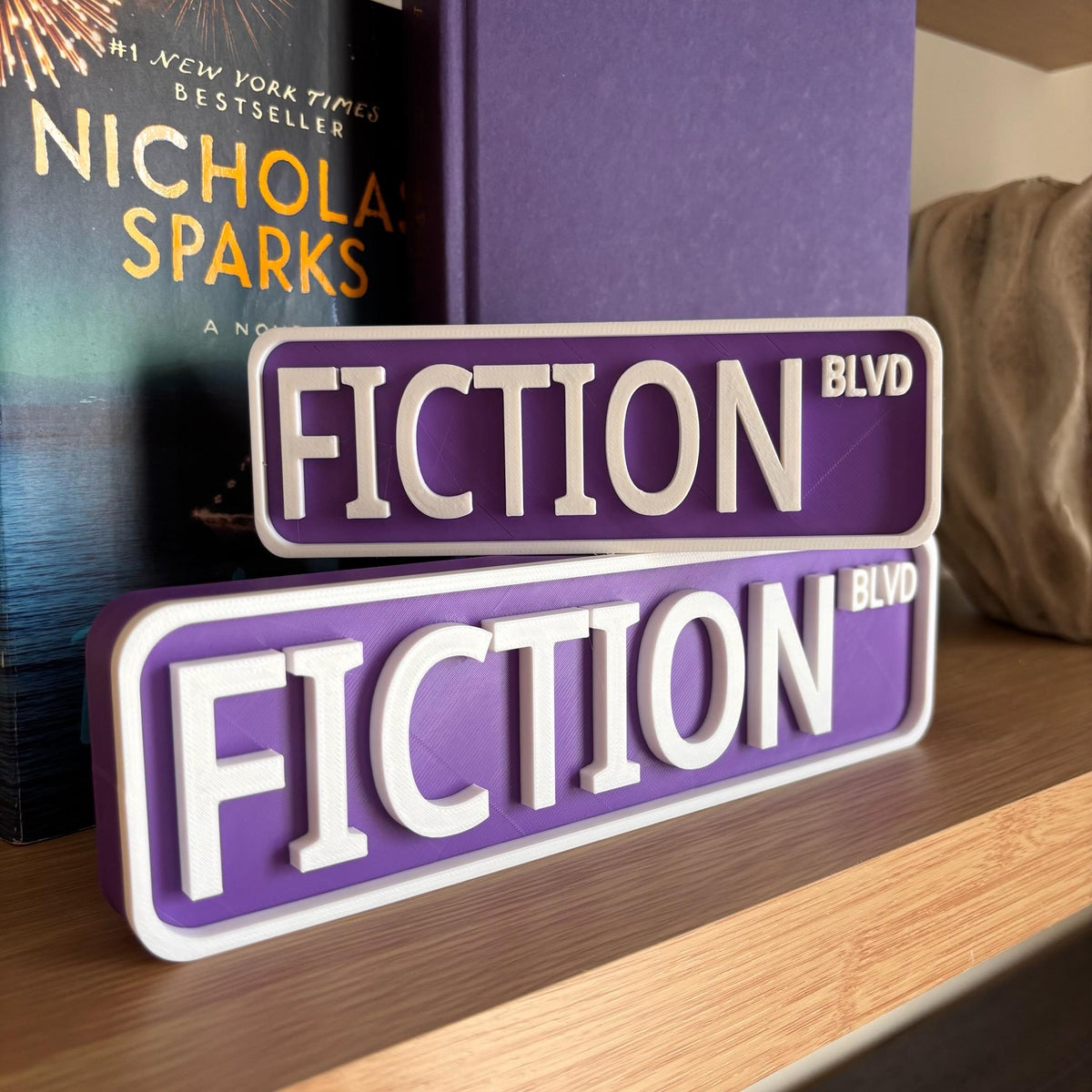 Purple and white FICTION BLVD street signs, small stacked on large, on a bookshelf