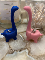Two dinosaur-shaped earring holders, one blue and one pink, with silver earrings on top of them. __etsy:6791612944
