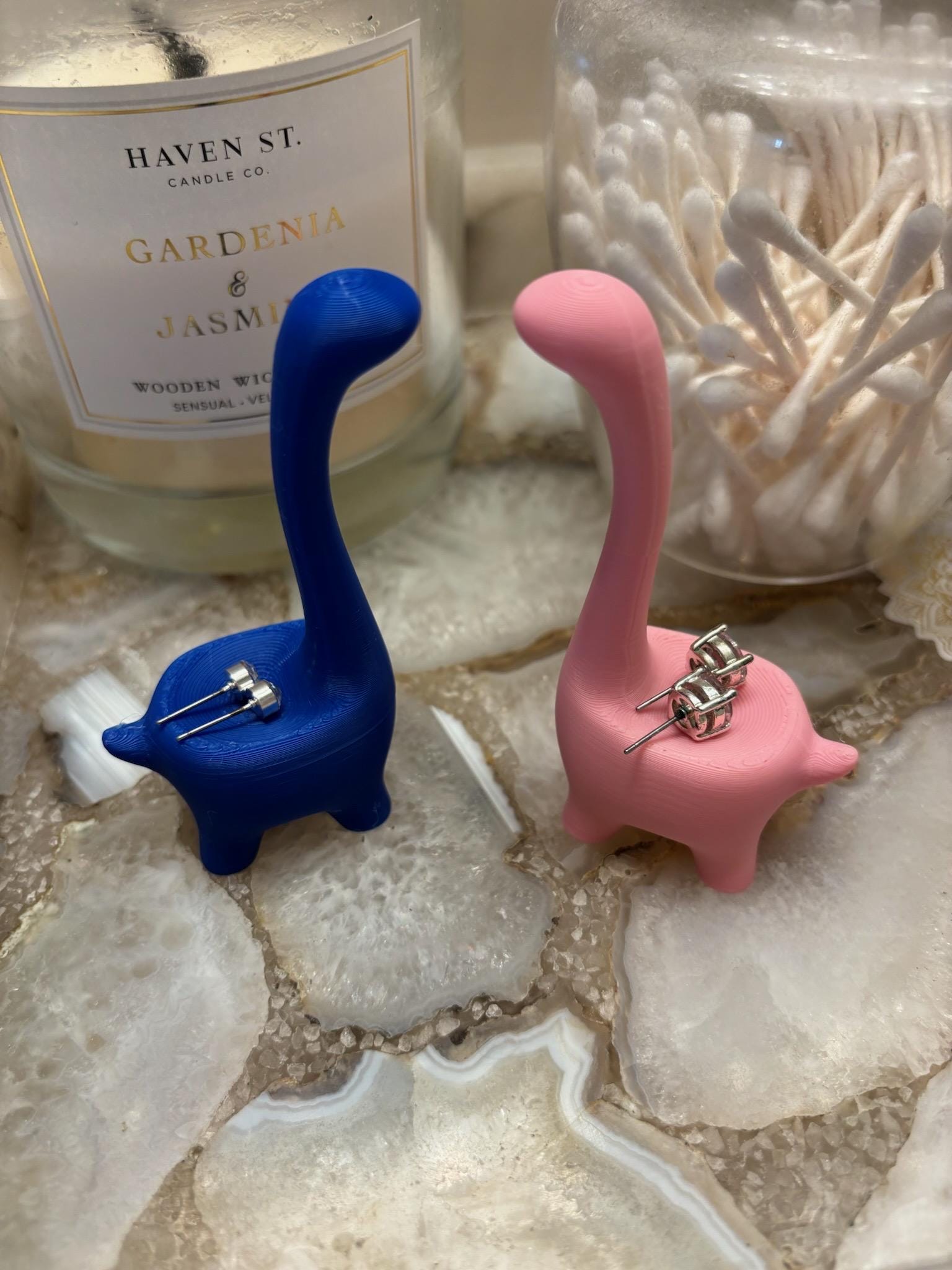 Two dinosaur shaped earring holders, one blue and one pink, with silver earrings on each holder. __etsy:6791612960