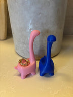 Two dinosaur-shaped ring holders, one pink and one blue. The pink dinosaur has a gold ring on its neck. __etsy:6839619431