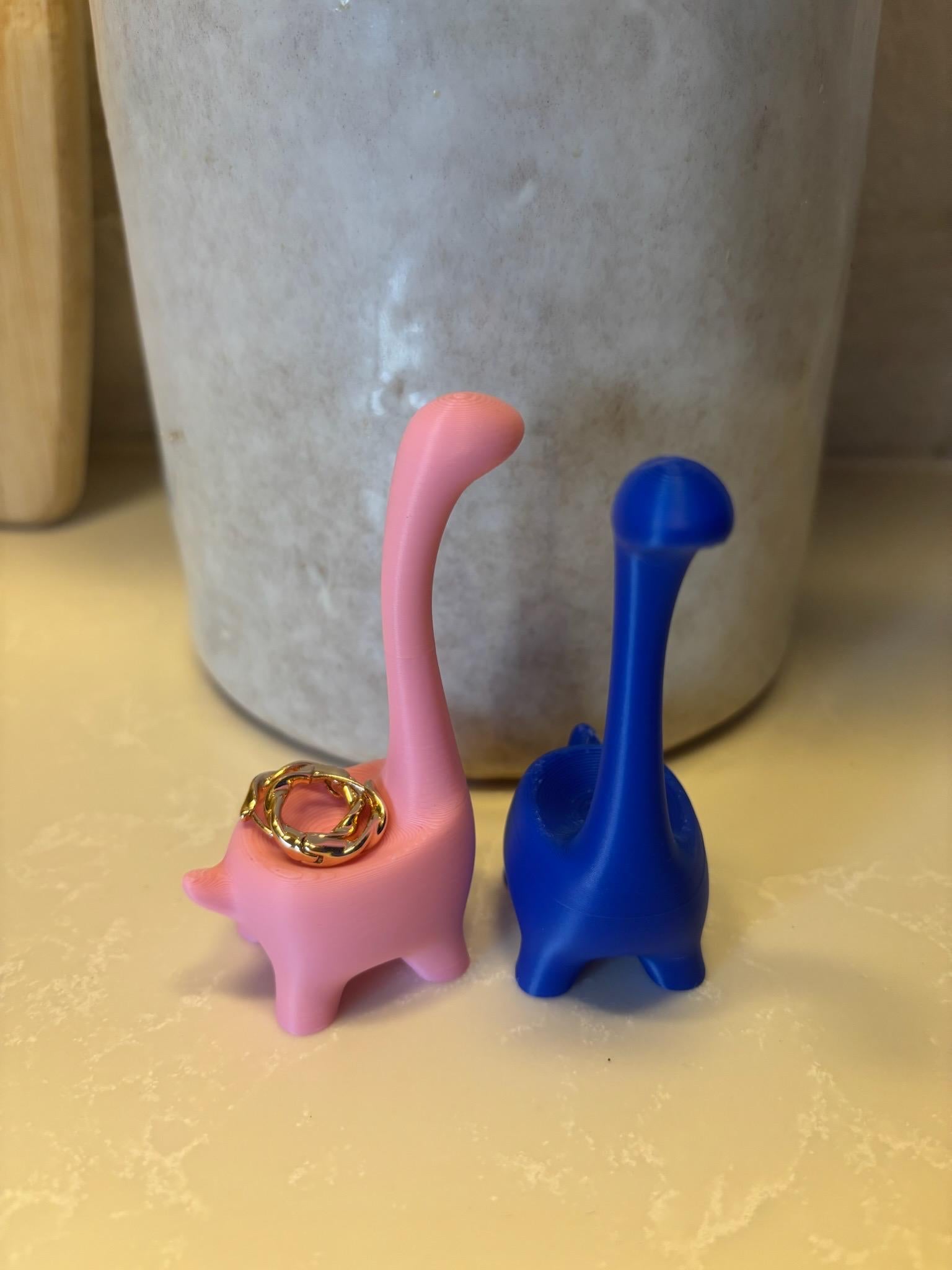 Two dinosaur-shaped ring holders, one pink and one blue. The pink dinosaur has a gold ring on its neck. __etsy:6839619431