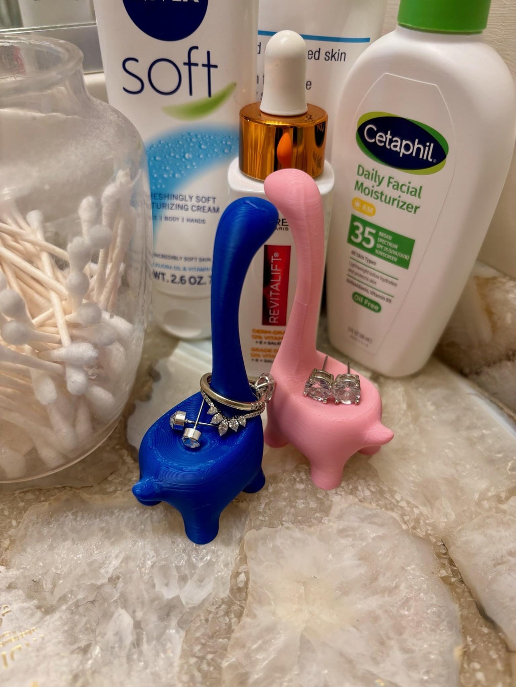 Two blue and pink dinosaur-shaped ring holders. The blue dinosaur is holding a ring and a pair of earrings. The pink dinosaur is holding a pair of earrings. The dinosaurs are sitting on a marble countertop. __etsy:6839619447