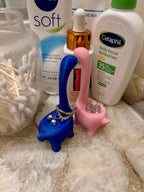 Two blue and pink dinosaur-shaped ring holders. The blue dinosaur is holding a ring and a pair of earrings. The pink dinosaur is holding a pair of earrings. The dinosaurs are sitting on a marble countertop. __etsy:6839619447