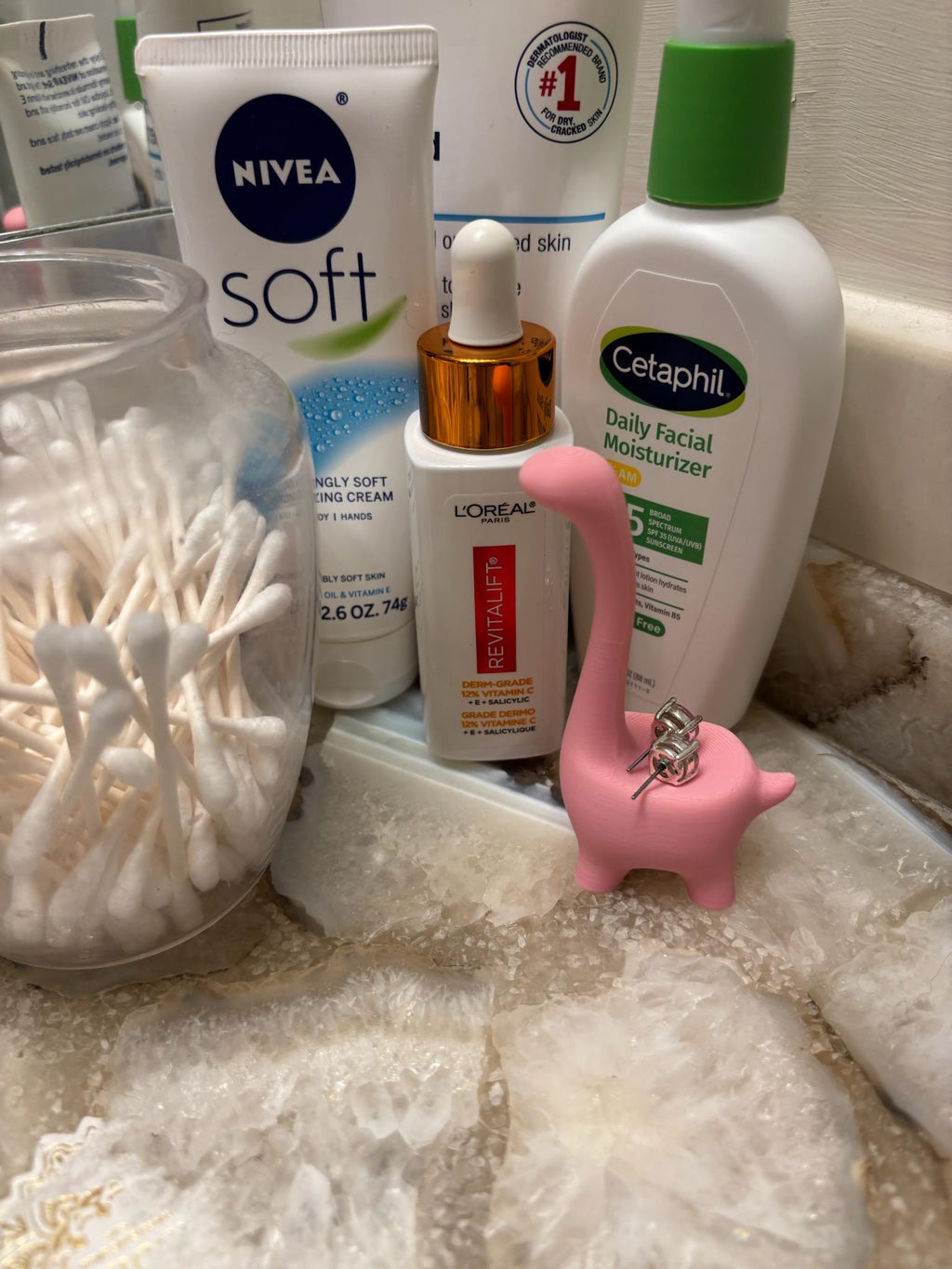 A pink dinosaur figurine with two silver earrings on its back. The figurine is on a white and gray surface in a bathroom. __etsy:6839619453