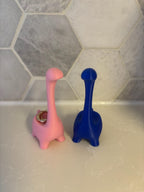 Two dinosaur-shaped ring holders, one pink and one blue, standing upright. The pink dinosaur has a gold ring on its back __etsy:6839622193