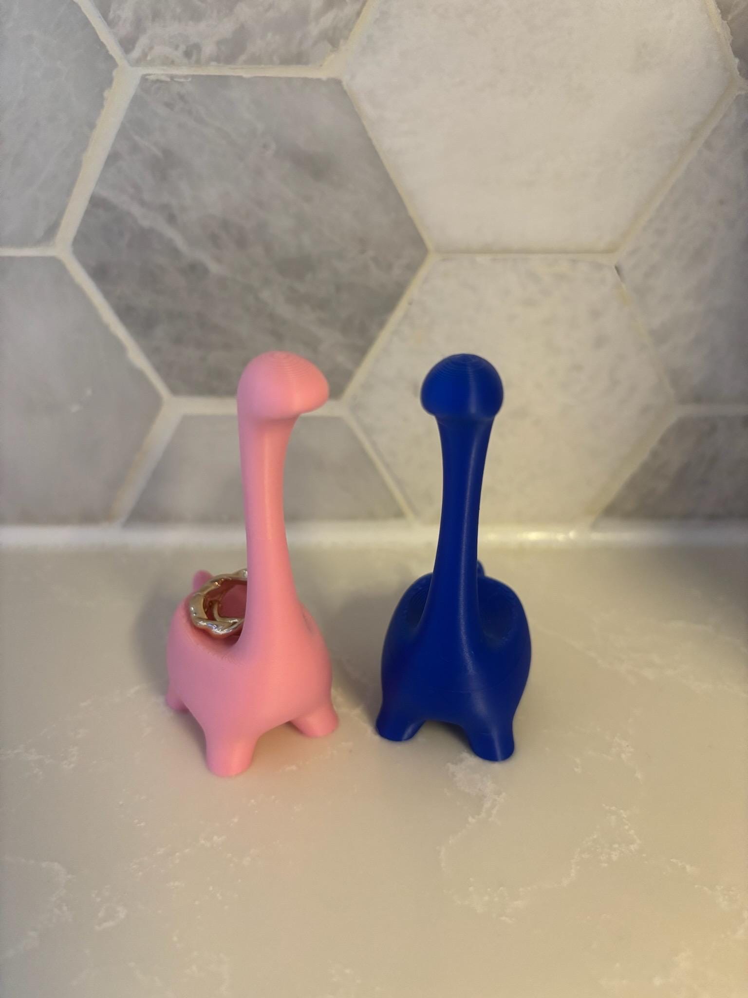 Two dinosaur-shaped ring holders, one pink and one blue, standing upright. The pink dinosaur has a gold ring on its back __etsy:6839622193