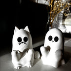 Two small white figurines sit on a surface: one shaped like a cat with round eyes and whiskers, and one shaped like a ghost with round eyes and a small smile. A vase with dried flowers is blurred in the background. __etsy:7159226238
