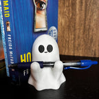A small smiling white ghost figurine holding a pen with black eyes in front of a book titled The Housemaid by Freida McFadden, against a dark wooden background. __etsy:7159226252