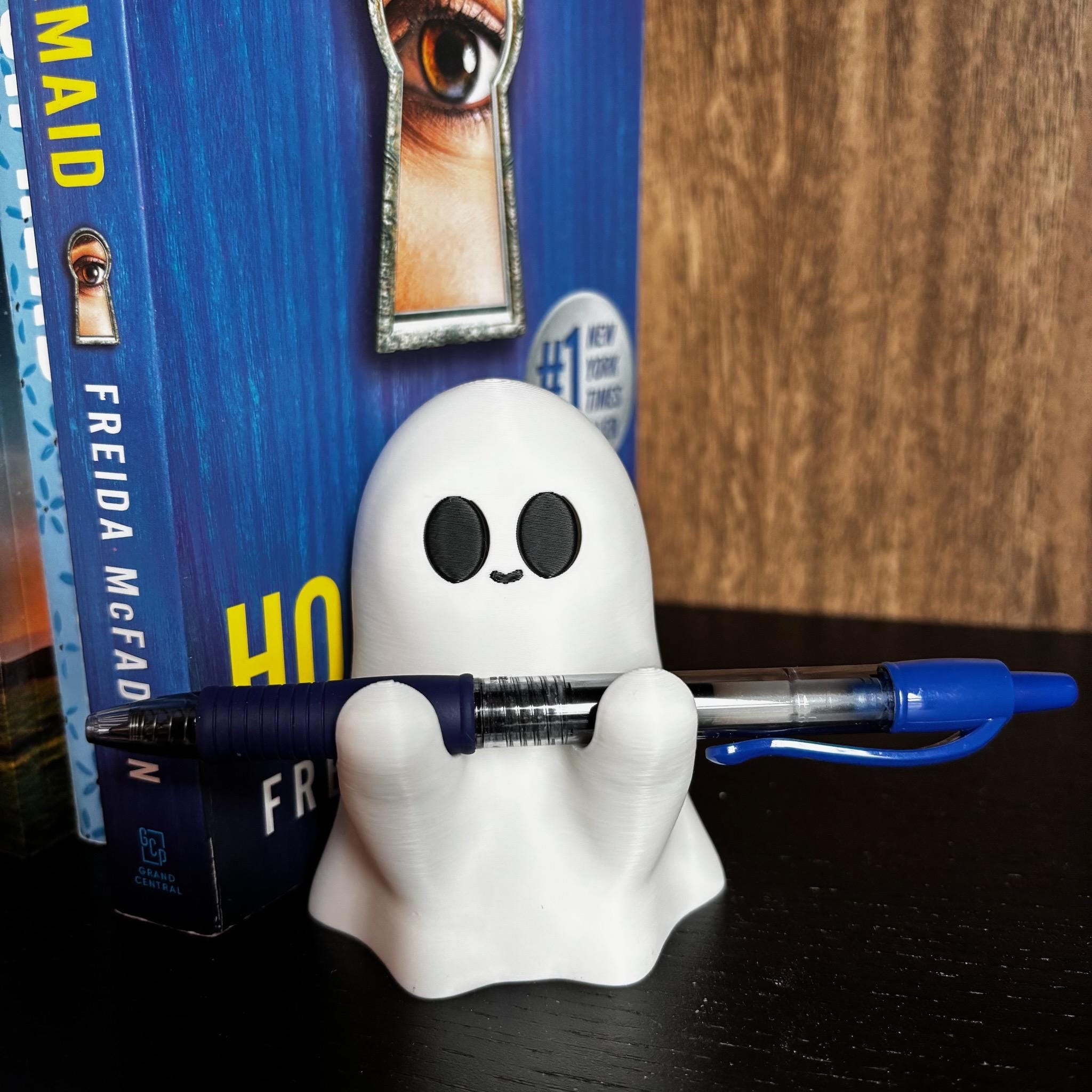 A small smiling white ghost figurine holding a pen with black eyes in front of a book titled The Housemaid by Freida McFadden, against a dark wooden background. __etsy:7159226252