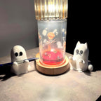 Two small white figurines, one a ghost holding a pen and the other a cat, sit beside a lamp with a red liquid base, casting warm light on a flat surface in an otherwise dimly lit room. __etsy:7159280490