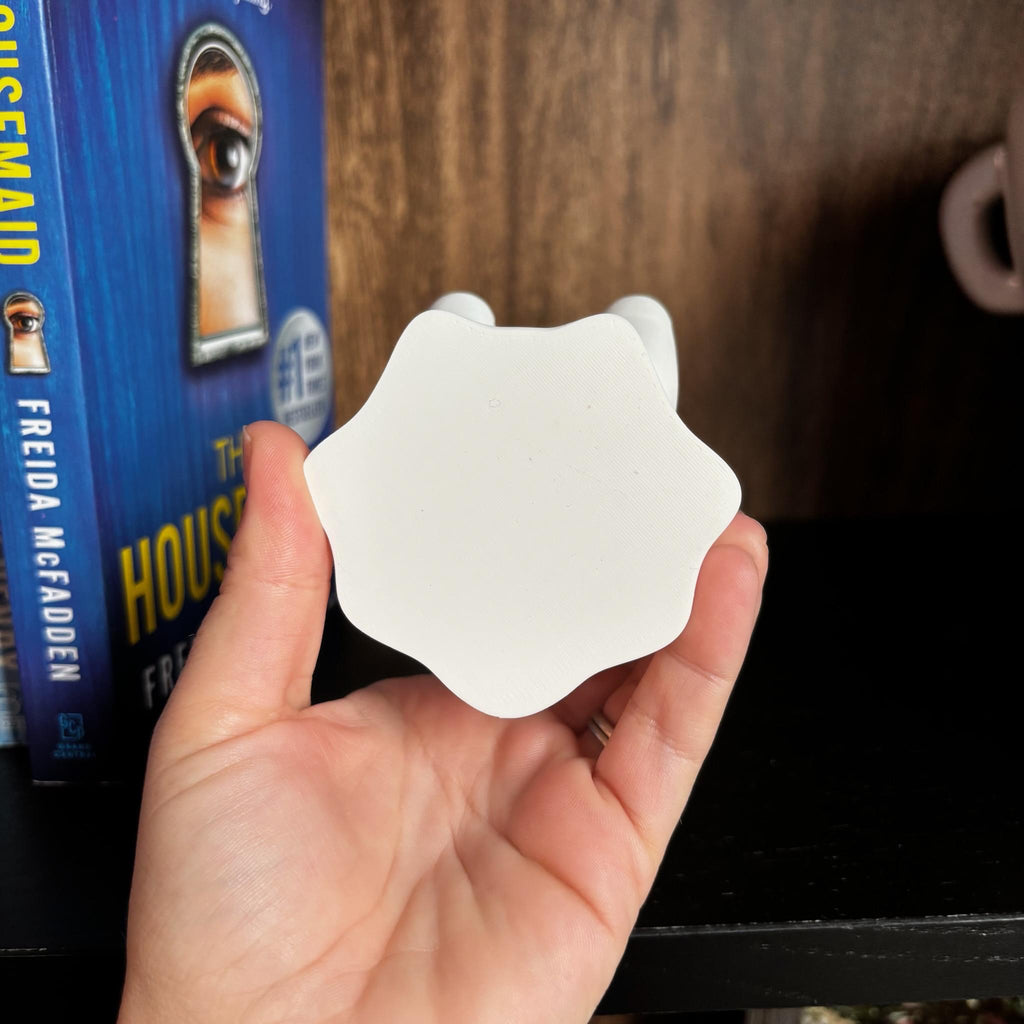 A hand holds the bottom of a 3d printed ghost with a flower-like base in front of a shelf. Behind it is a book titled The Housemaid by Freida McFadden, with a keyhole and eye on the cover. __etsy:7207215731